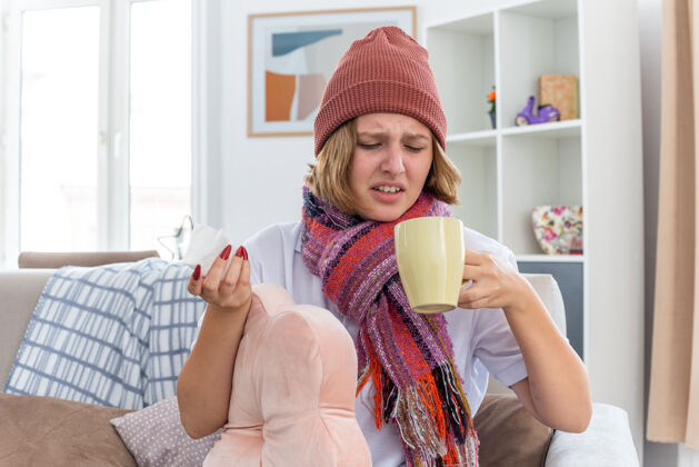 病不健康的年轻女子戴着暖和的帽子 戴着围巾 看起来不舒服 生病了 手里拿着一杯热茶和患感冒和流感的纸巾 坐在明亮的客厅里的椅子上持有生活椅子