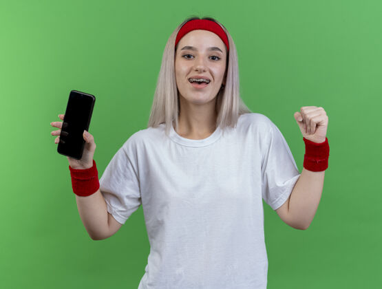拳头快乐的年轻白人运动女孩戴着背带 戴着头带和腕带 握紧拳头 拿着电话快乐举行运动