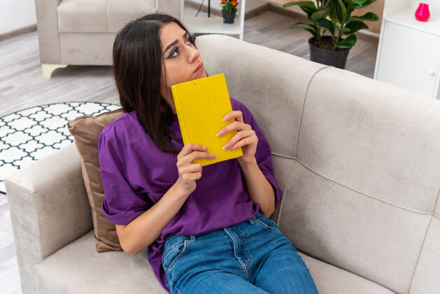 女人一个穿着休闲服的年轻女孩拿着一本书 困惑地坐在客厅的沙发上生活坐着一旁