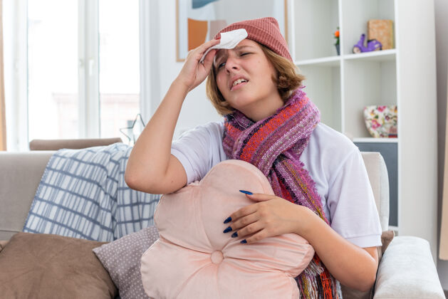 健康不健康的年轻女子戴着暖和的帽子 戴着围巾 看起来不舒服 摸着额头 发烧 头痛 患感冒和流感 坐在明亮的客厅沙发上沙发暖和感冒
