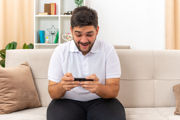 年轻一个穿着休闲服的年轻人拿着智能手机玩游戏 快乐而愉快地坐在客厅里的沙发上男人房间手机