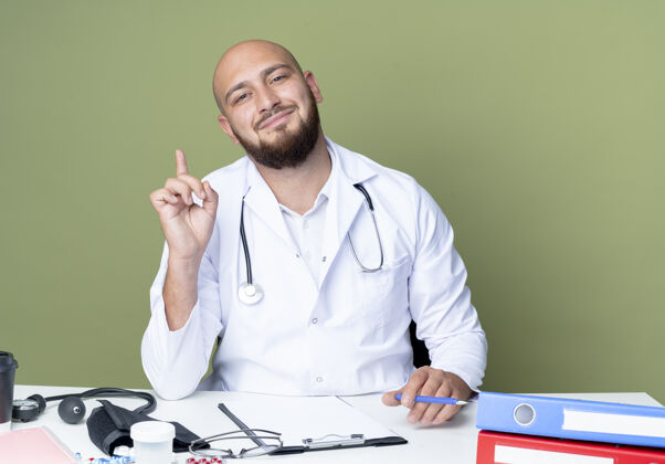 绿色高兴的年轻秃头男医生穿着医用长袍和听诊器坐在办公桌旁工作办公桌医生