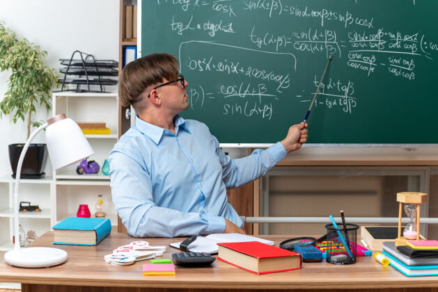 笔记年轻的男老师戴着眼镜在教室里讲课 自信地坐在课桌旁 黑板前放着书和笔记数学课学校