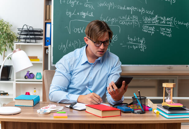笔记年轻的男老师戴着眼镜看着他的智能手机 自信地坐在课桌旁 教室的黑板前放着书和笔记坐着前面黑板