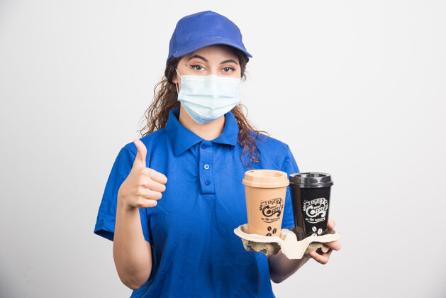 手势身穿蓝色制服 戴着医用面罩的女人 手里拿着两杯咖啡 在白色的脸上摆出ok的姿势工人两个