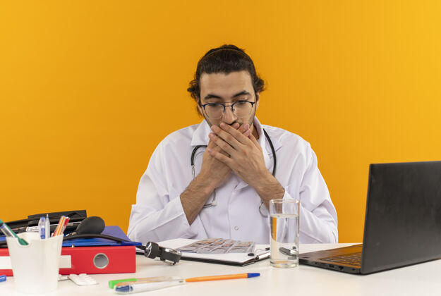 穿戴着医用眼镜 穿着医用长袍 带听诊器的年轻男医生坐在办公桌旁 吓了一跳长袍复制黄色