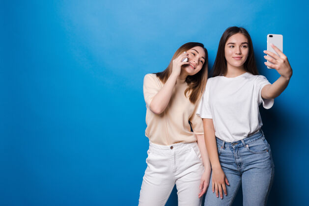 画像两个漂亮的女孩在蓝色的墙上自拍乐趣欢呼青年