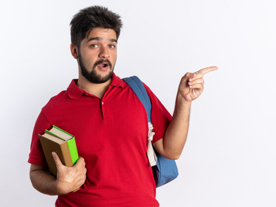 拿着年轻的留着胡子的学生 穿着红色马球衫 背着书包 拿着笔记本 看上去既高兴又惊讶 用食指指着旁边站着壁板惊喜指着