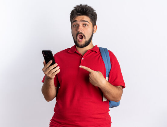 指着一个年轻的留着胡子的学生 穿着红色马球衫 背着背包 拿着智能手机 用食指指着它 站在那里显得很惊讶站着背包胡子