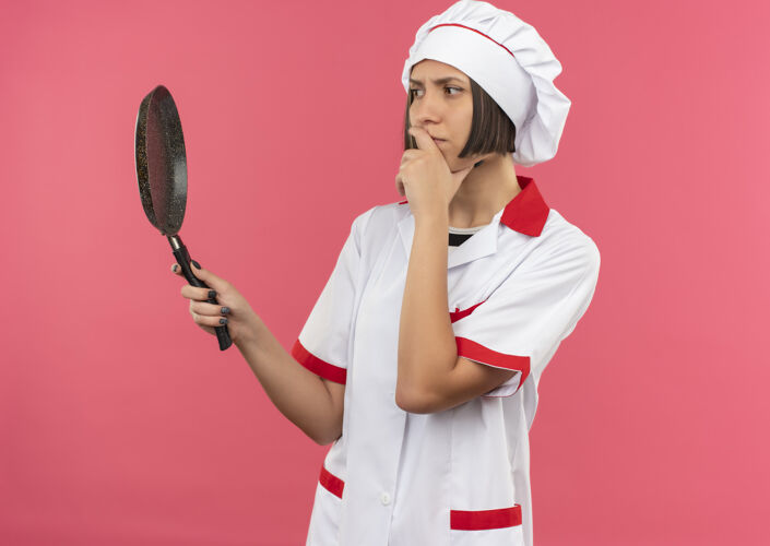 下巴体贴的年轻女厨师 穿着厨师制服 手放在下巴上 手举着手 看着粉红色墙上孤立的煎锅烹饪粉色锅