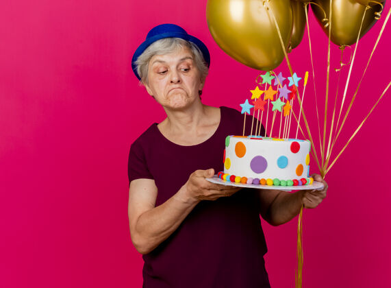 穿困惑的老妇人戴着聚会帽 手里拿着氦气球 看着粉红色的生日蛋糕老人困惑粉色