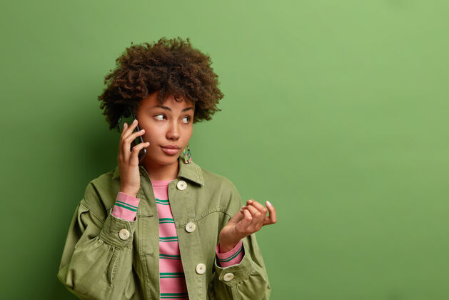 衣服体贴的年轻美国黑人妇女举手打电话考虑从对话者那里听到的信息穿着时髦的衣服隔着鲜艳的绿色墙壁设备思维肖像
