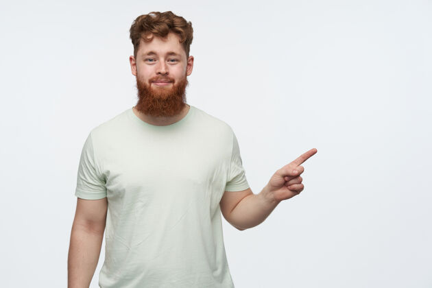 外观年轻的留着胡子的红发男人的肖像 穿着一件空白的t恤 用手指着右边的复印空间好帅气室内