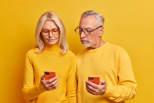 阅读当代老年夫妇自信科技用户专注于智能手机检查信息戴眼镜矫正视力隔离在黄色的工作室墙上老年人男性退休
