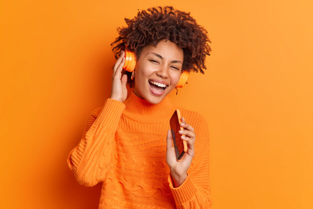 舞蹈快乐的美国黑人女模特唱着歌 把智能手机放在嘴边 就好像麦克风戴着无线耳机 微笑着隔着鲜艳的橙色墙壁美丽女性无线