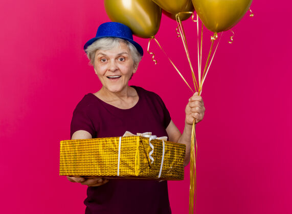 举行惊讶的老妇人戴着派对帽 手里拿着氦气球和粉红色的礼盒惊喜帽子女人