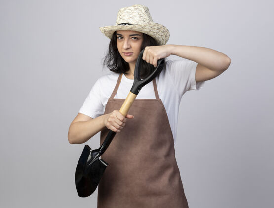 年轻自信的年轻黑发女园丁穿着制服 戴着园艺帽 手里拿着一把铁锹自信白色园艺