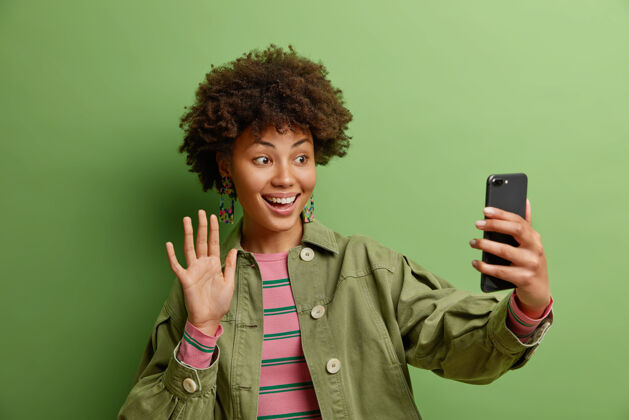 快乐科技和在线生活方式的概念微笑的女人说嗨喜欢视频通话使用高速互联网连接穿着时尚夹克隔离在绿色的墙壁上通信手机夹克