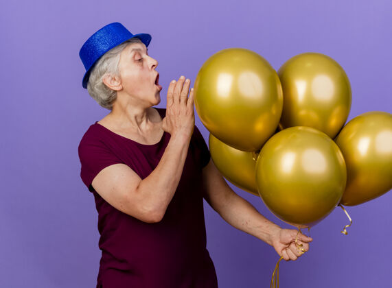 惊喜惊讶的老妇人戴着聚会的帽子站在紫色的气球上看着氦气球帽子氦女人