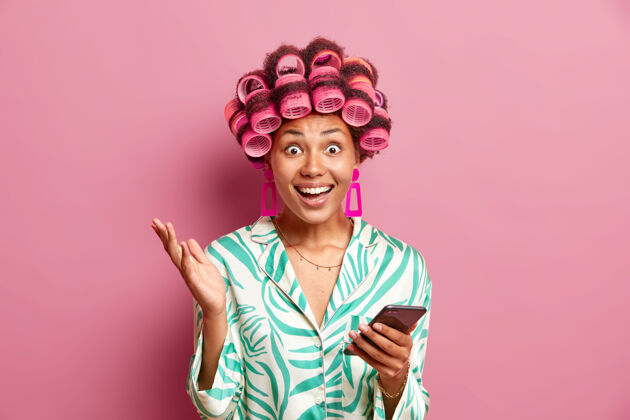 请穿着丝绸睡衣的积极女人使用手机下载新应用程序使卷发与粉红墙隔离辊姿势兴奋互联网