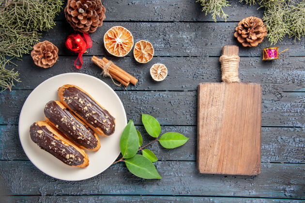 早餐俯视图：椭圆形盘子上的巧克力蛋糕 圣诞玩具 冷杉树叶 肉桂 干橙子和砧板 放在深色的木质地面上顶部橘子枞树