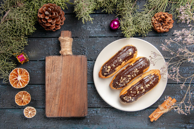 深色俯视图巧克力eclairs在白色椭圆形板冷杉树枝和锥圣诞玩具干花枝干桔子肉桂和砧板在深色木桌上干的木板枞树
