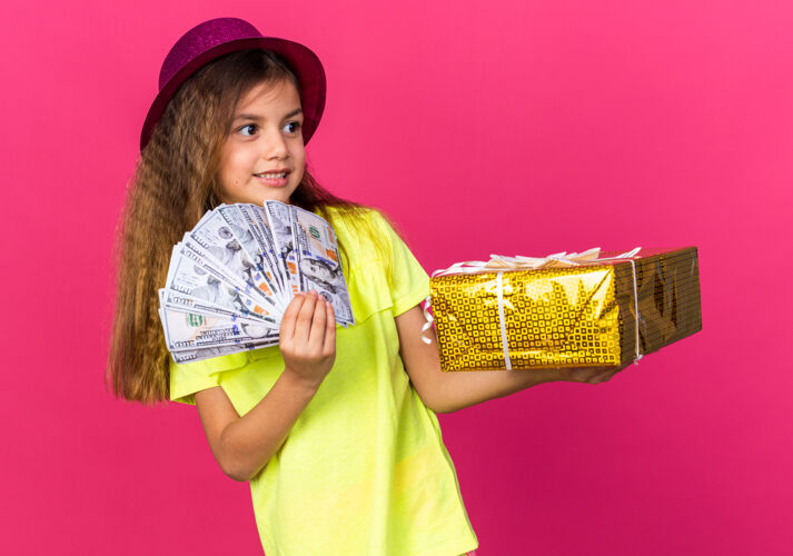 白种人可爱的白人小女孩 戴着紫色派对帽 手里拿着礼盒和钱 看着粉色墙壁上与世隔绝的一面 还有复制空间女孩孤立小
