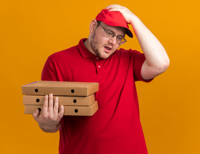 头焦急的超重的年轻送货员戴着眼镜 手里拿着披萨盒 手放在头上 隔离在橙色的墙上 留着复印空间孤立焦虑年轻