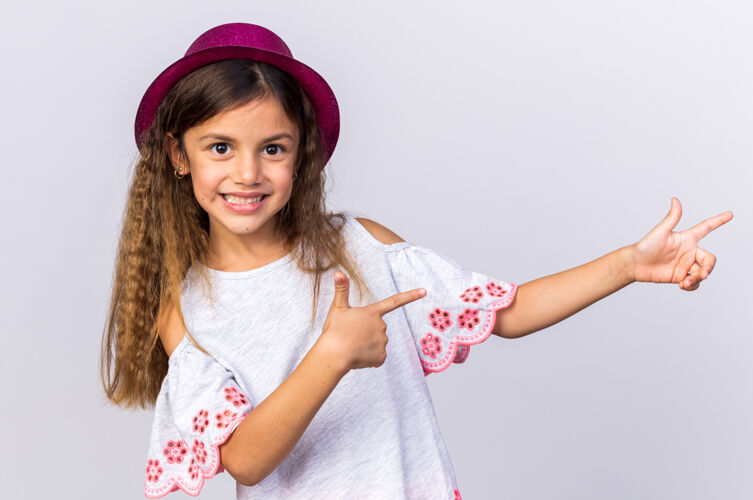 白种人面带微笑的白人小女孩 戴着紫色派对帽 指着隔离在白色墙壁上的一面 留着复制空间女孩小微笑
