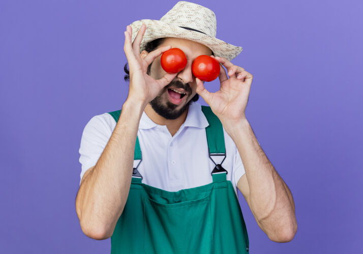 年轻年轻的留着胡须的园丁 穿着连体衣 戴着帽子 手里拿着新鲜的西红柿 眼睛蒙着 站在蓝色的墙上微笑着穿西红柿花园