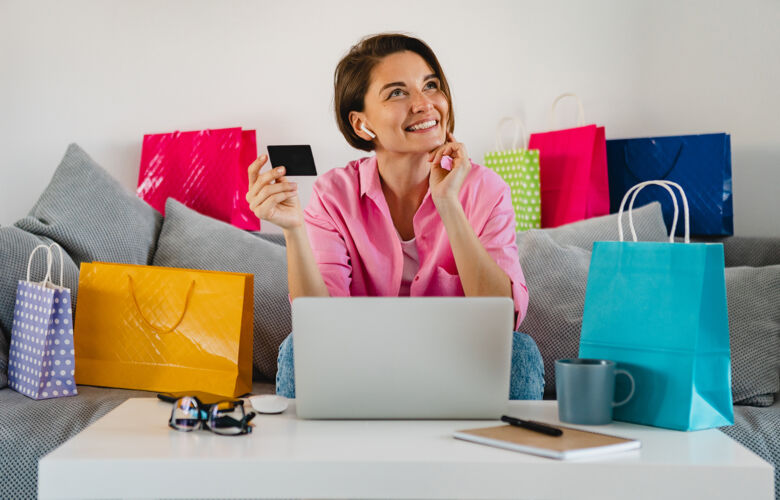 笔记本电脑在家里的沙发上 穿着粉色衬衫的快乐微笑的女人在五颜六色的购物袋中拿着信用卡 在笔记本电脑上在线支付房子沙发支付
