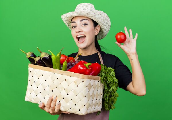 兴奋年轻漂亮的女园丁围着围裙 戴着帽子 手里拿着装满蔬菜的篮子 站在绿色的墙边 表现出西红柿的快乐和兴奋站着女人抱着