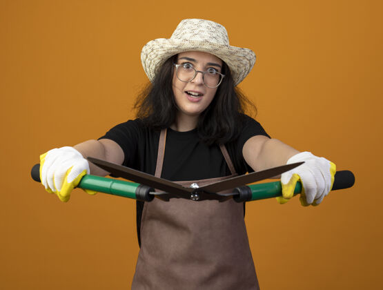 黑发惊讶的是 年轻的深色皮肤的女园丁戴着眼镜 穿着制服 戴着园艺帽和手套 把园艺剪孤立在橙色的墙上眼镜眼镜手套