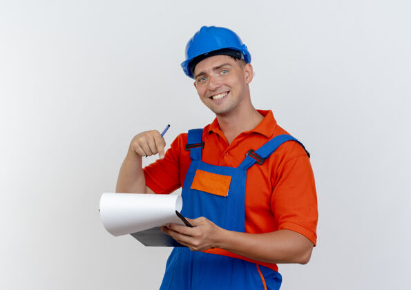 微笑微笑的年轻男性建筑工人穿着制服 戴着安全帽 指着剪贴板背景点年轻
