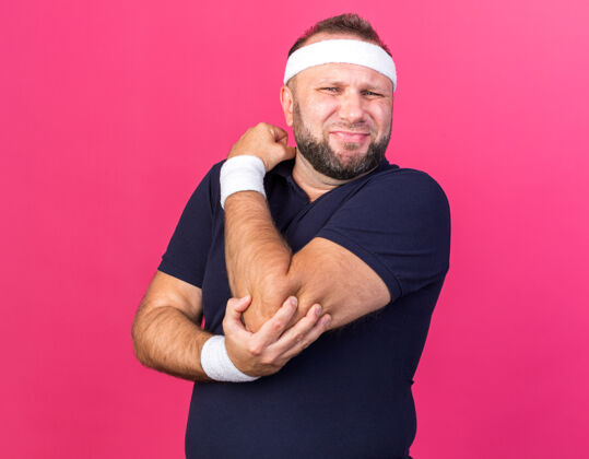 抱着疼痛的成年斯拉夫运动型男子戴着头带和腕带 手肘孤立地放在粉色墙壁上 留有复制空间戴男人肘部