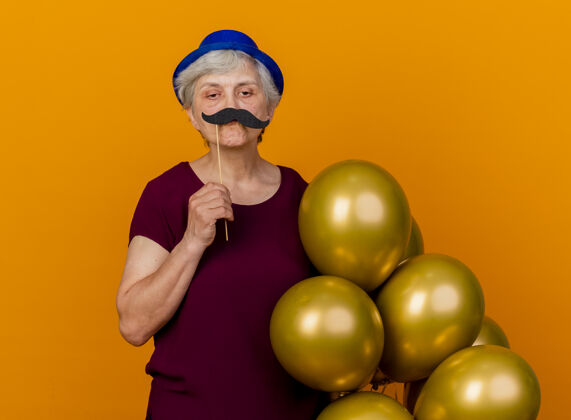 氦自信的老妇人戴着派对帽 手里拿着氦气球和假胡子 贴在橙色的墙上胡子人举行