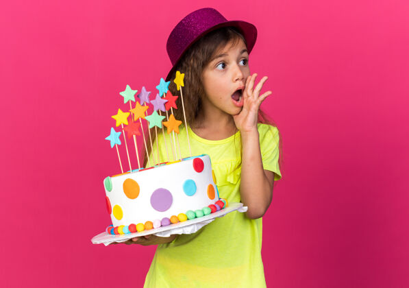 壁板震惊的小白种女孩 戴着紫色派对帽 手放在脸上 拿着生日蛋糕 看着粉色墙壁上孤立的一面 还有复制空间小白种人手