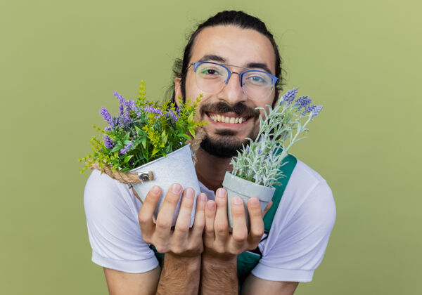 人年轻的留着胡须的园丁身穿连体衣 手里拿着盆栽植物 站在浅绿色的墙上 兴高采烈地微笑着看着前方姿势表情花园