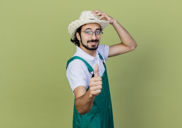人年轻的留着胡须的园丁 穿着连体衣 戴着帽子 站在浅绿色的墙上 微笑着竖起大拇指 看着前面表演连身衣感觉