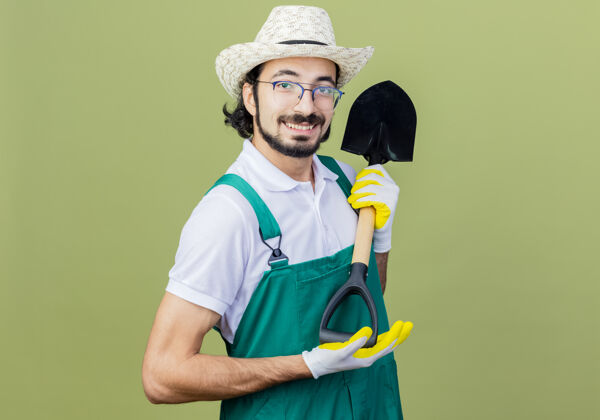 铲子年轻的留着胡须的园丁 穿着连体衣 戴着帽子 站在浅绿色的墙上微笑着看着前方感觉穿着展示