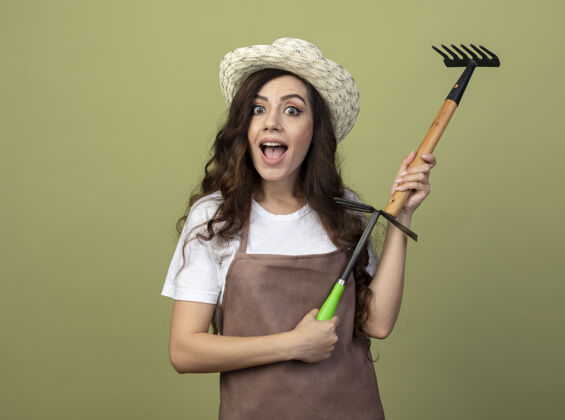 花园兴奋的年轻女园丁穿着制服 戴着园艺帽 把耙子和锄头耙子隔离在橄榄绿的墙上人人穿