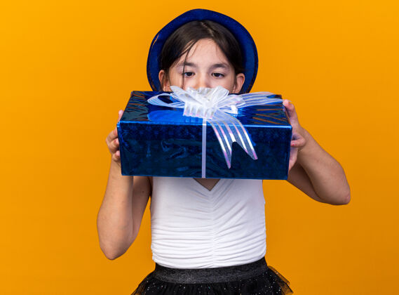 女孩兴奋的年轻白种人女孩拿着蓝色派对帽 看着孤立在橙色墙上的礼品盒 还有复制空间孤立兴奋年轻