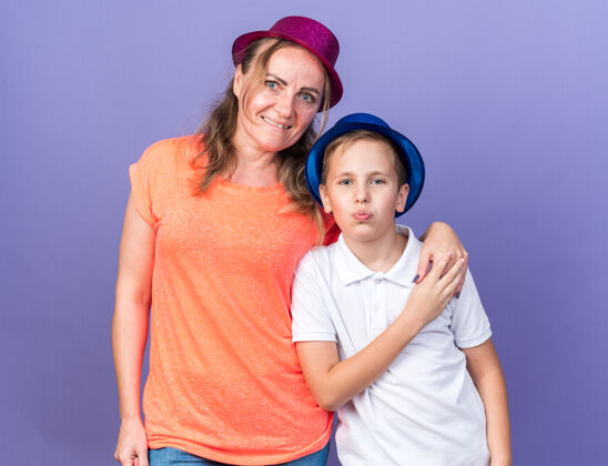 拜托高兴的年轻斯拉夫男孩戴着蓝色的聚会帽站在他的母亲戴着紫色的聚会帽孤立在紫色墙上与复制空间斯拉夫人男孩孤立