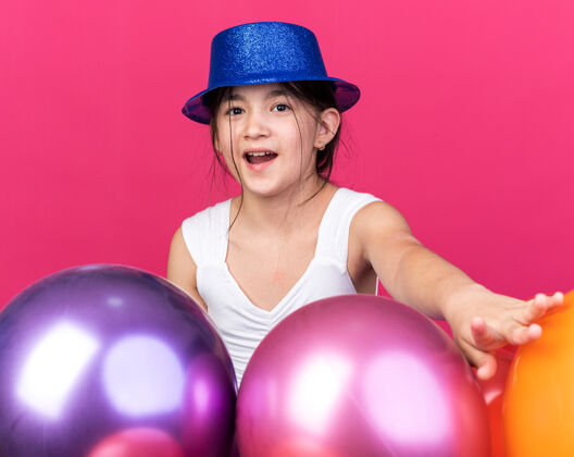气球惊讶的年轻白人女孩戴着蓝色派对帽站在粉红色的墙上 氦气球与复制空间隔离白种人女孩惊喜