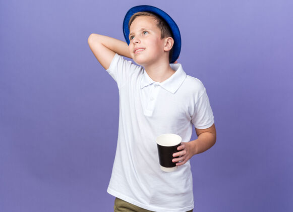 头面带微笑的斯拉夫男孩 戴着蓝色派对帽 手放在头上 手拿纸杯 看着紫色墙上孤立的一面 还有复制空间壁板帽子孤立