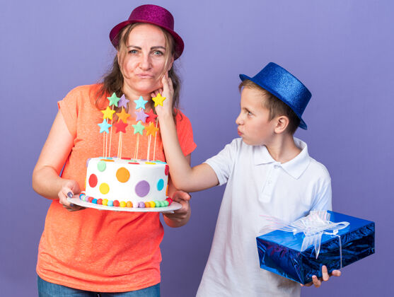 抱着高兴的年轻斯拉夫男孩戴着蓝色派对帽拿着礼盒拉着妈妈的脸颊戴着紫色派对帽拿着生日蛋糕隔离在紫色的墙上有复制空间斯拉夫人男孩脸颊
