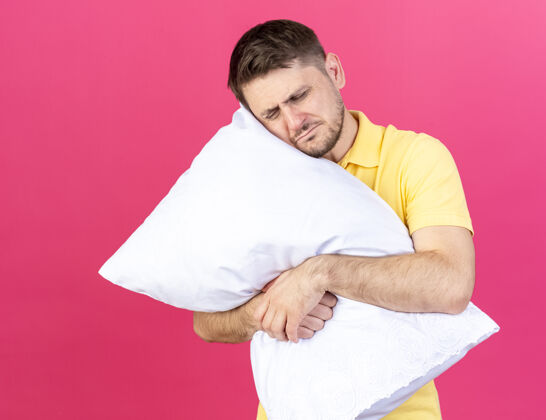 脸疼痛的年轻金发病夫拥抱并把头放在隔离在粉红色墙上的枕头上疾病男人人