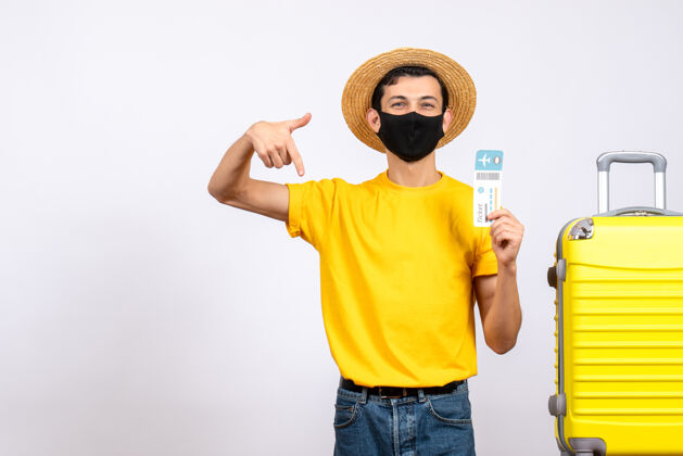 黄色正面图：戴着草帽和口罩的年轻人站在拿着旅行票的黄色手提箱旁抱着手提箱站着