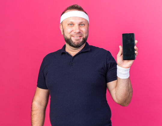 斯拉夫面带微笑的成年斯拉夫运动型男子戴着头带和腕带 手持电话 隔离在粉色墙壁上 留有复印空间微笑运动手持