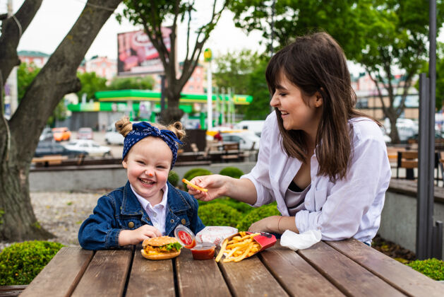 汉堡快乐可爱的小女孩坐在她的母亲与汉堡和薯条在一个咖啡馆的桌子上小女孩休闲孩子